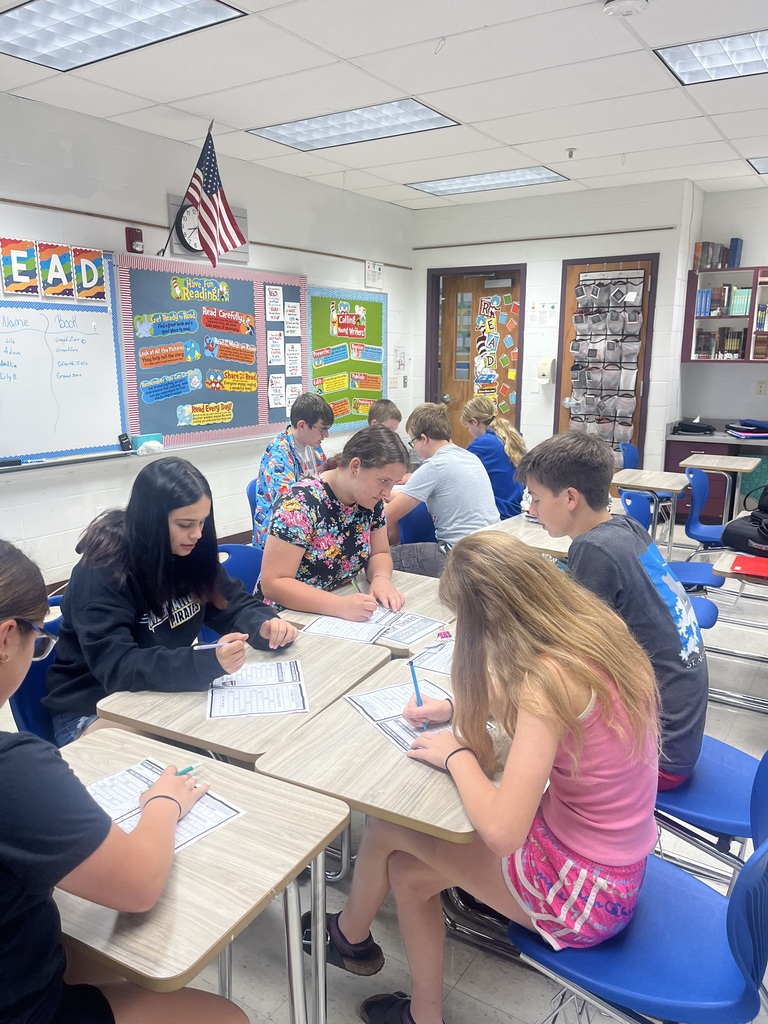 Students are sitting with their desks facing each other. Students are holing game questions. They are working together to find the clue.