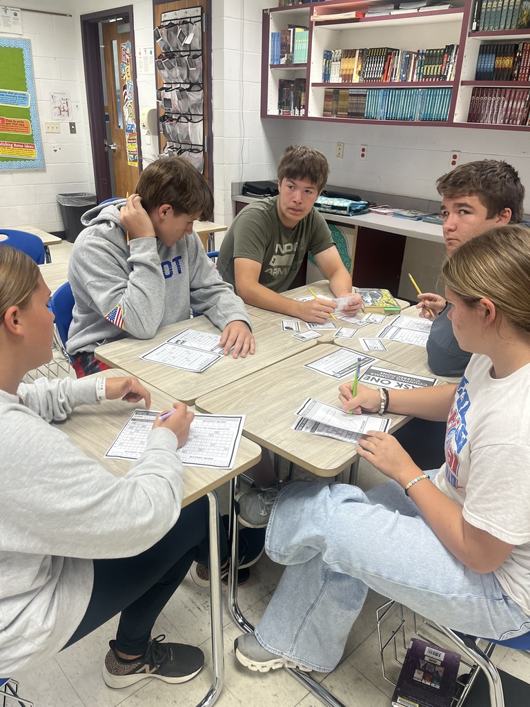 Students are sitting with their desks facing each other. Students are holing game questions. They are working together to find the clue.