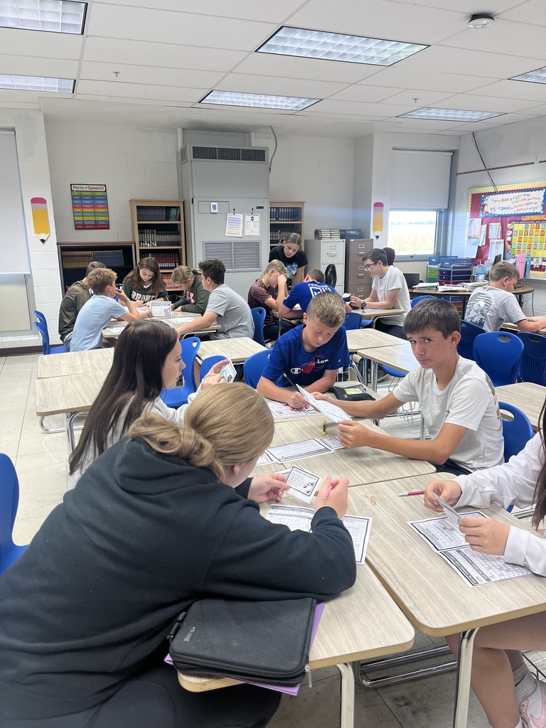 Students are sitting with their desks facing each other. Students are holing game questions. They are working together to find the clue.