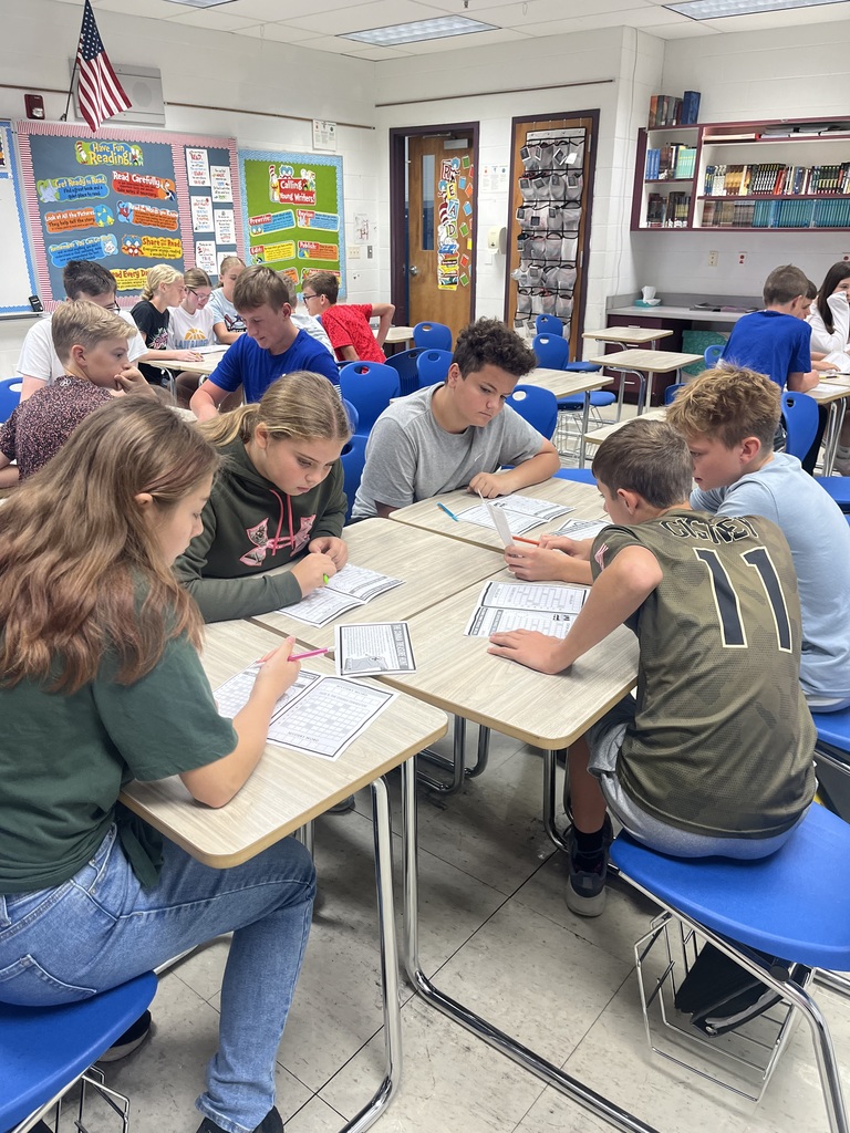Students are sitting with their desks facing each other. Students are holing game questions. They are working together to find the clue.