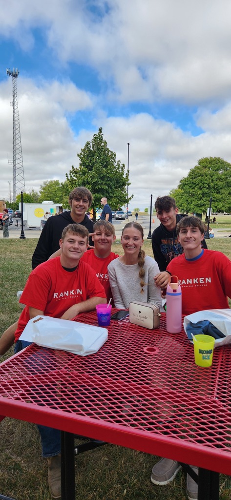 Six students are sitting at a red picnic table. They are posing together for the picture.