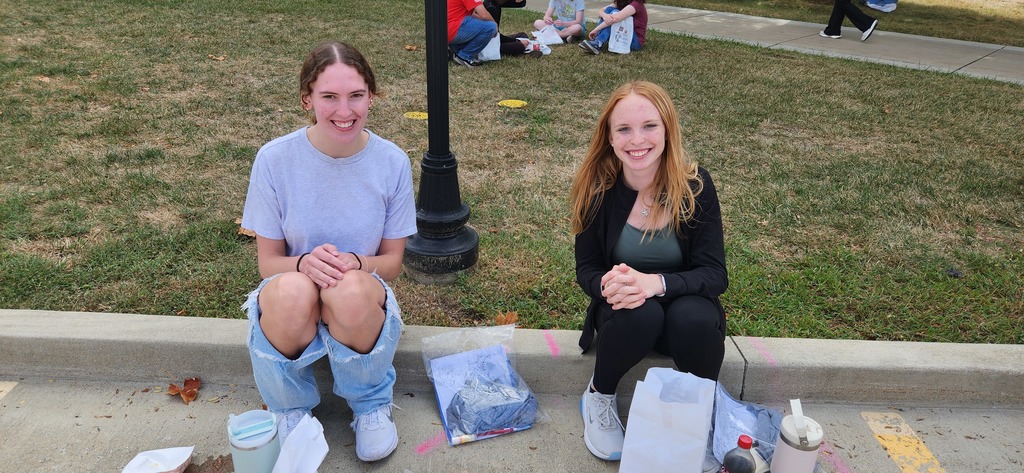 Two students are sitting together on a curb. They are visiting after lunch.