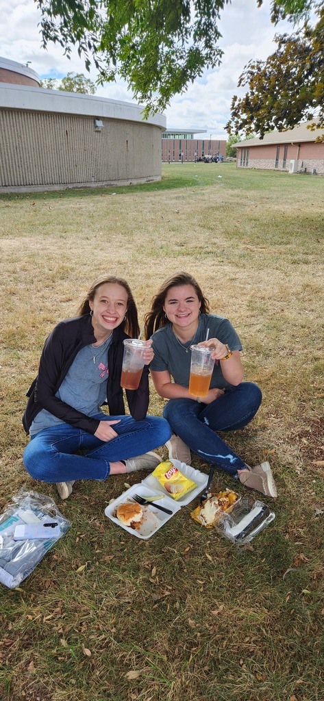 Two students are sitting together on the grass. They are drinking teas they purchased and eating thier lunch.