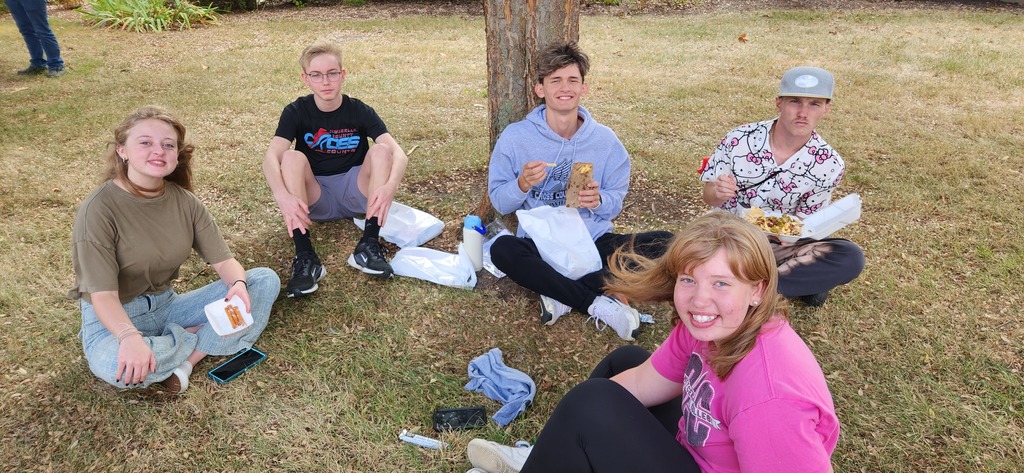 Students are sitting in a circle on the ground. They are eating their lunches.