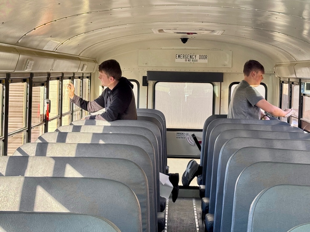 Two students are standing between seats of the bus on opposite sides. They are wiping the windows with a rag.
