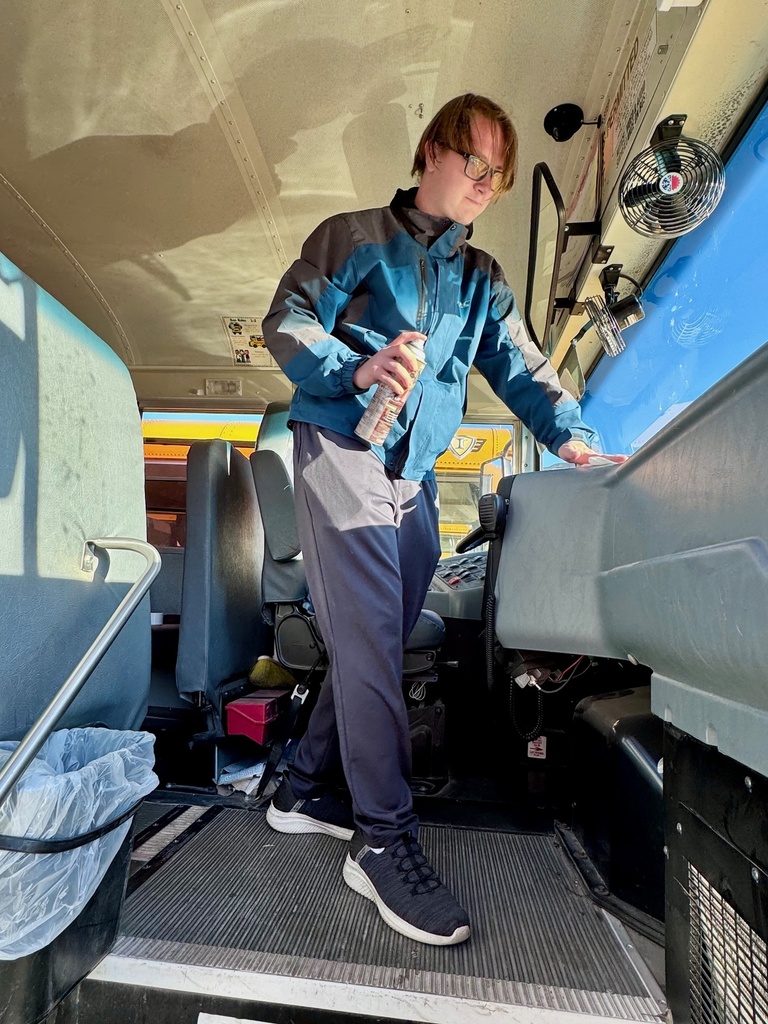 A student is standing in front of the driver's seat. He is wiping the dash and holding a can of spray.