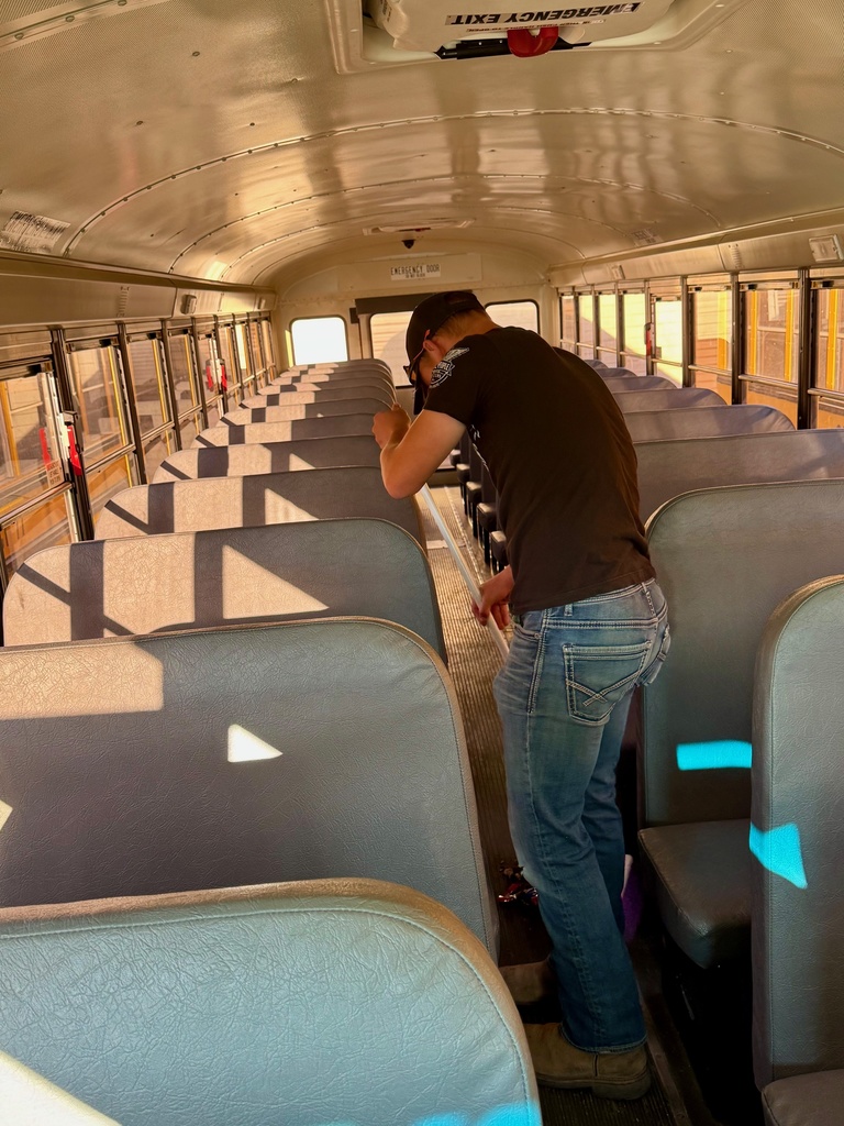A student is in the aisle of a bus. He is sweeping the floor.