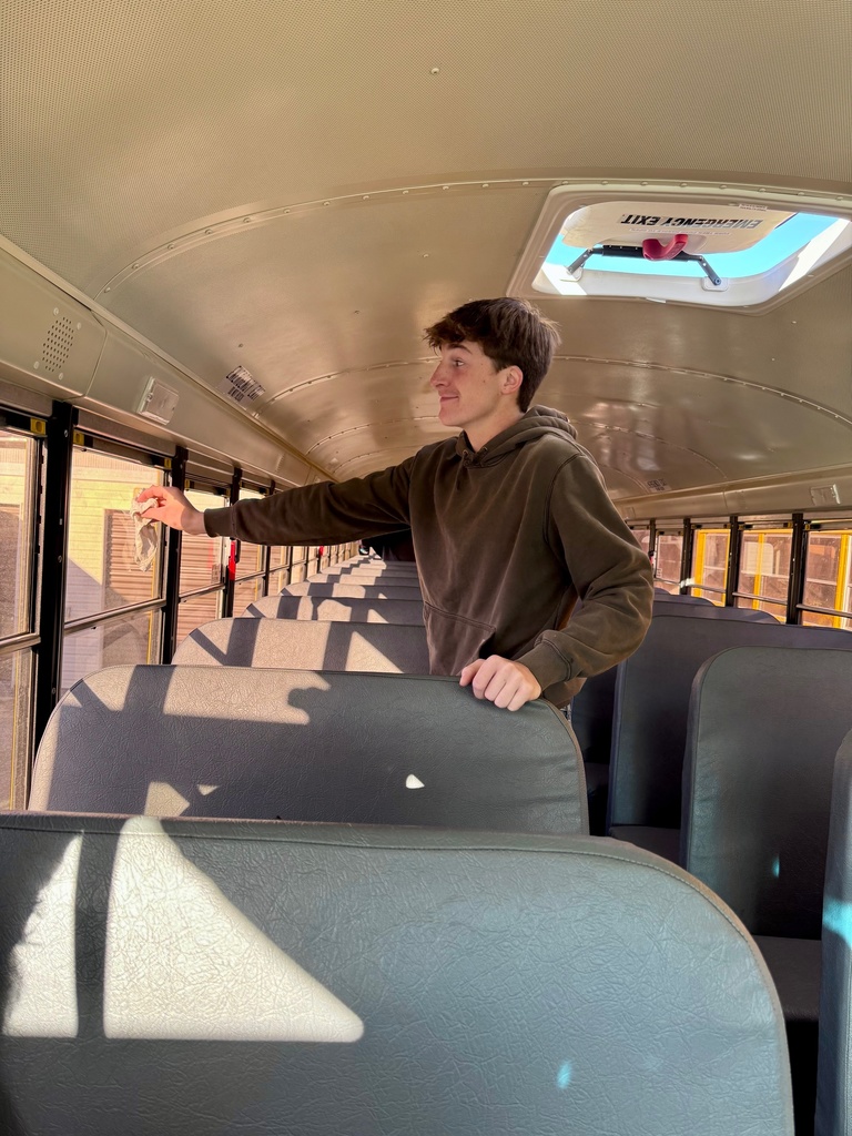 A student is standing between bus seats. He is wiping the windows with a rag.