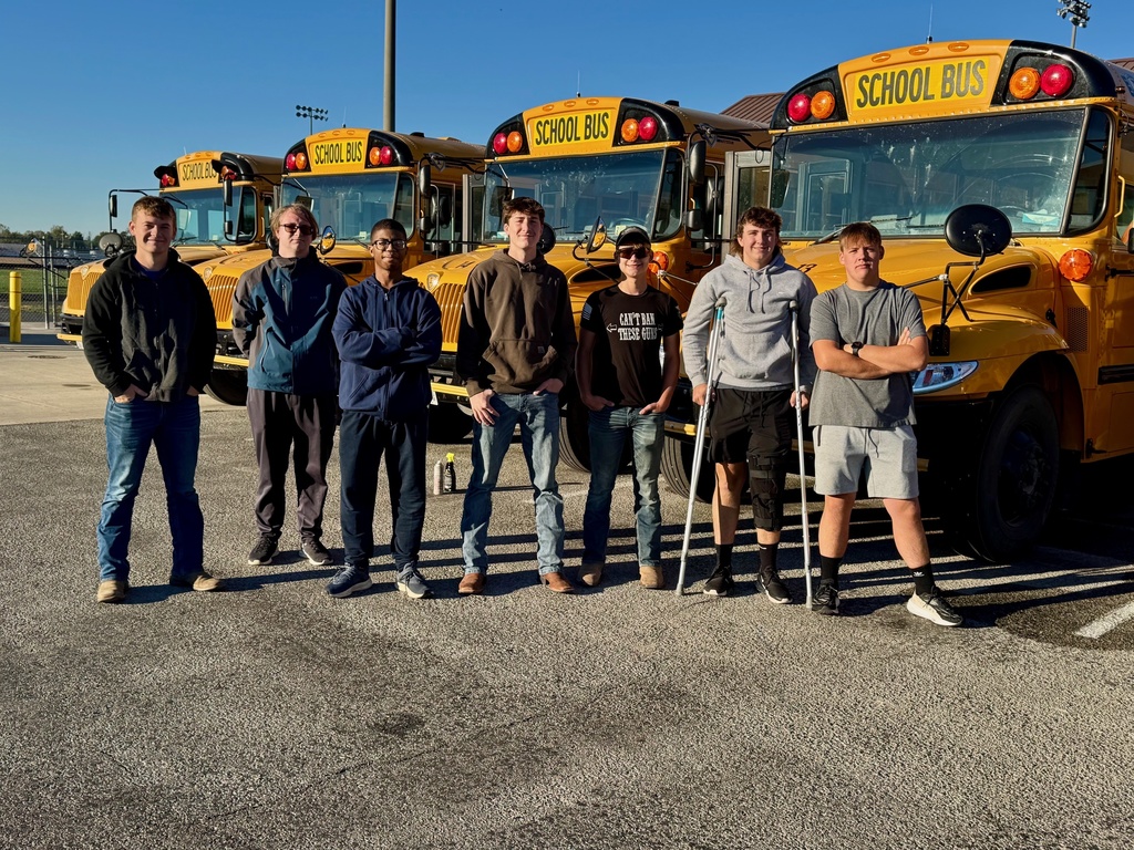 The seven students are standing side by side in front of the school buses.