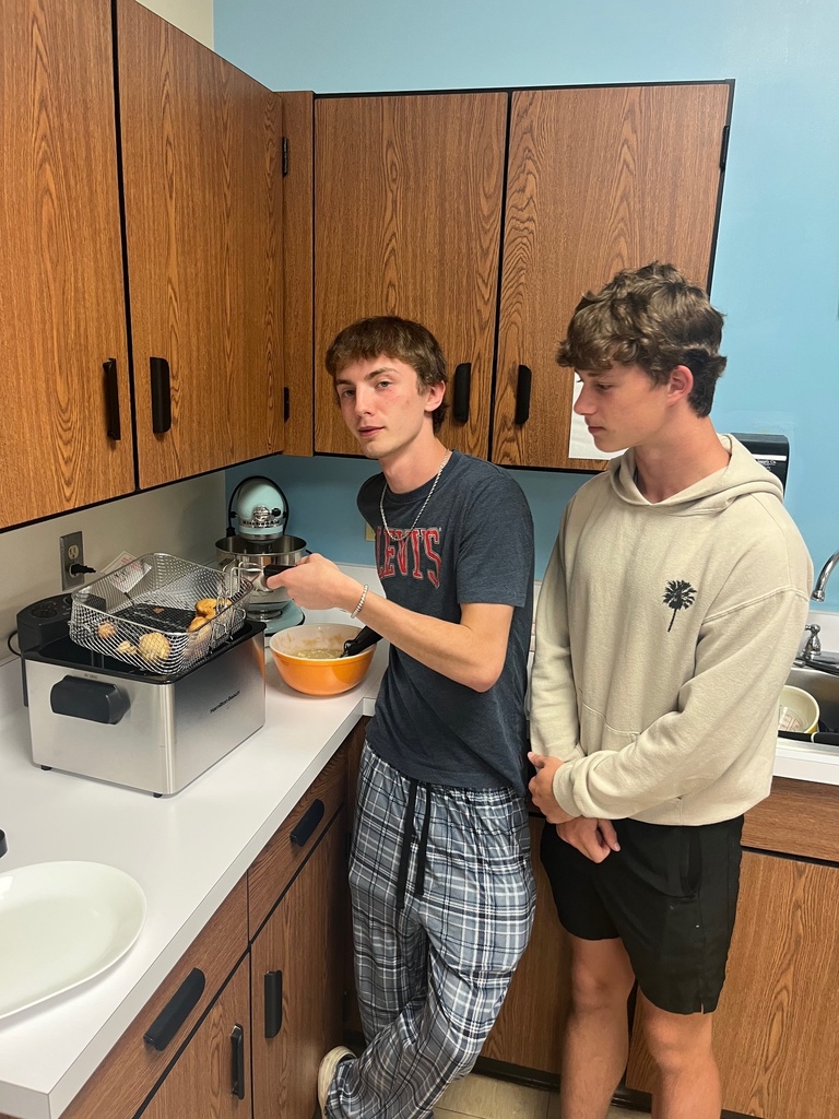 A student is pulling the basket out of the fryer. It has four apple fritters. Another student is looking on.