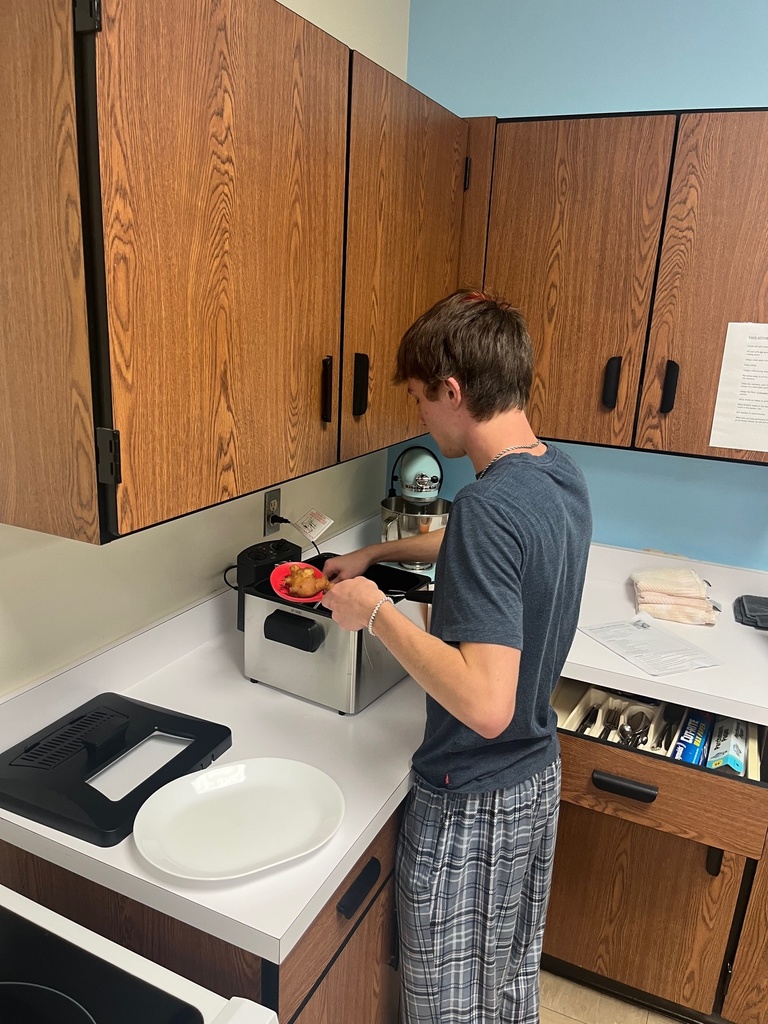A student is standing next to the counter with a red spoon in his hand. He is dishing up apple fritters from the fryer.