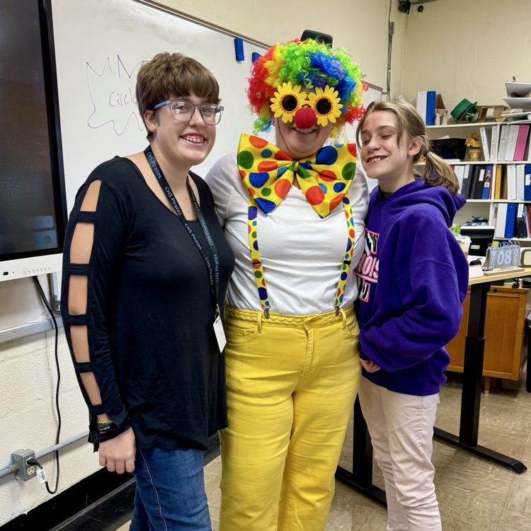 two students are standing next to their teacher who is dressed up like a clown. The teacher has a wig and a nose and sunglasses.
