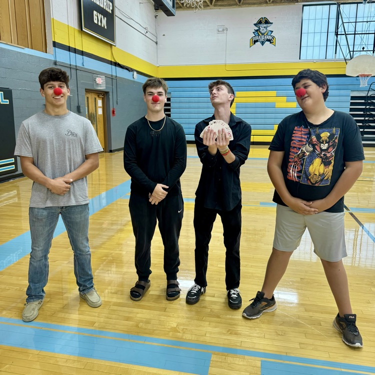 four students are standing side-by-side. three of them have clown noses in the fourth is holding a deck of cards.