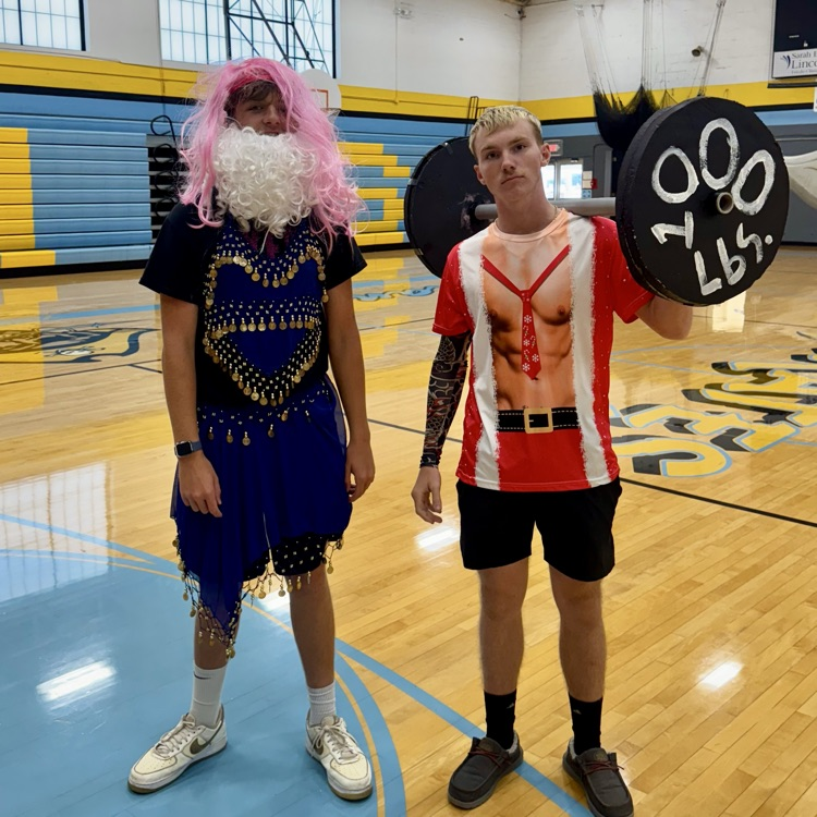 two students are standing side-by-side. One is stressed as a weight lifter. He is holding 1000 pound barbell. The other is dressed as a circus individual.