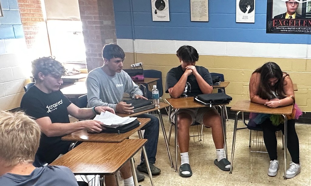 Three students are sitting at their desks. One is staring straight ahead. Another is flipping through paper. Another is resting his head on his hands.