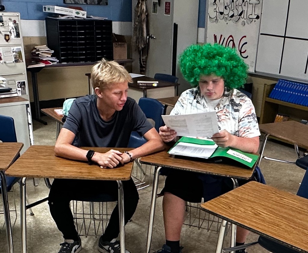 Two students are sitting at their desks. One is reading a packet. The other is looking at that packet.