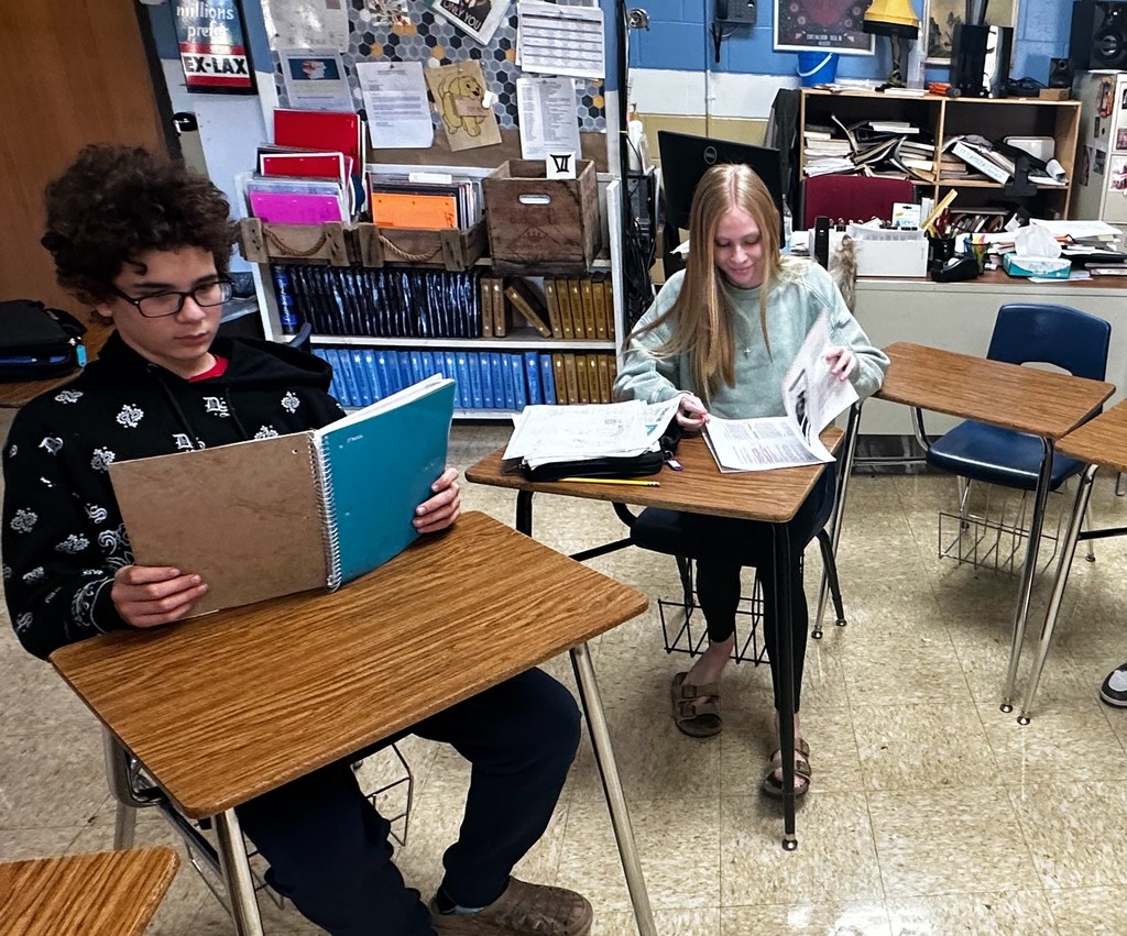 Two students are sitting at their desks. One is reading a notebook. Another is flipping through a packet.