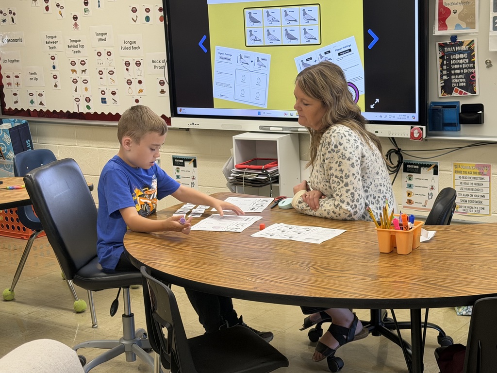 The student is holding a glue stick in his hand. He is looking over his paper. The teacher is looking on.