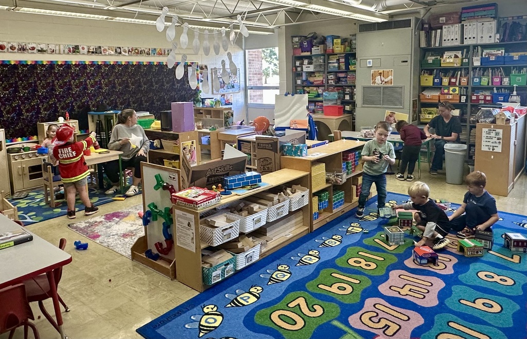 Students are playing in a preschool classroom. Two students are playing dress-up, while three are building a town on the floor.