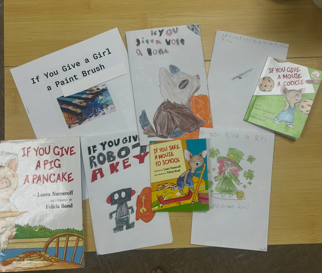 A tabletop display of children's books and handmade book covers inspired by Laura Numeroff's "If You Give..." series. Among the official titles like If You Give a Pig a Pancake, If You Give a Mouse a Cookie, and If You Take a Mouse to School, there are student-created versions with hand-drawn covers, including titles such as If You Give a Girl a Paint Brush, If You Give a Wolf a Bone, If You Give a Robot a Key, and If You Give a Girl a Leprechaun Hat. Each homemade book features colorful illustrations and playful interpretations of the original series.