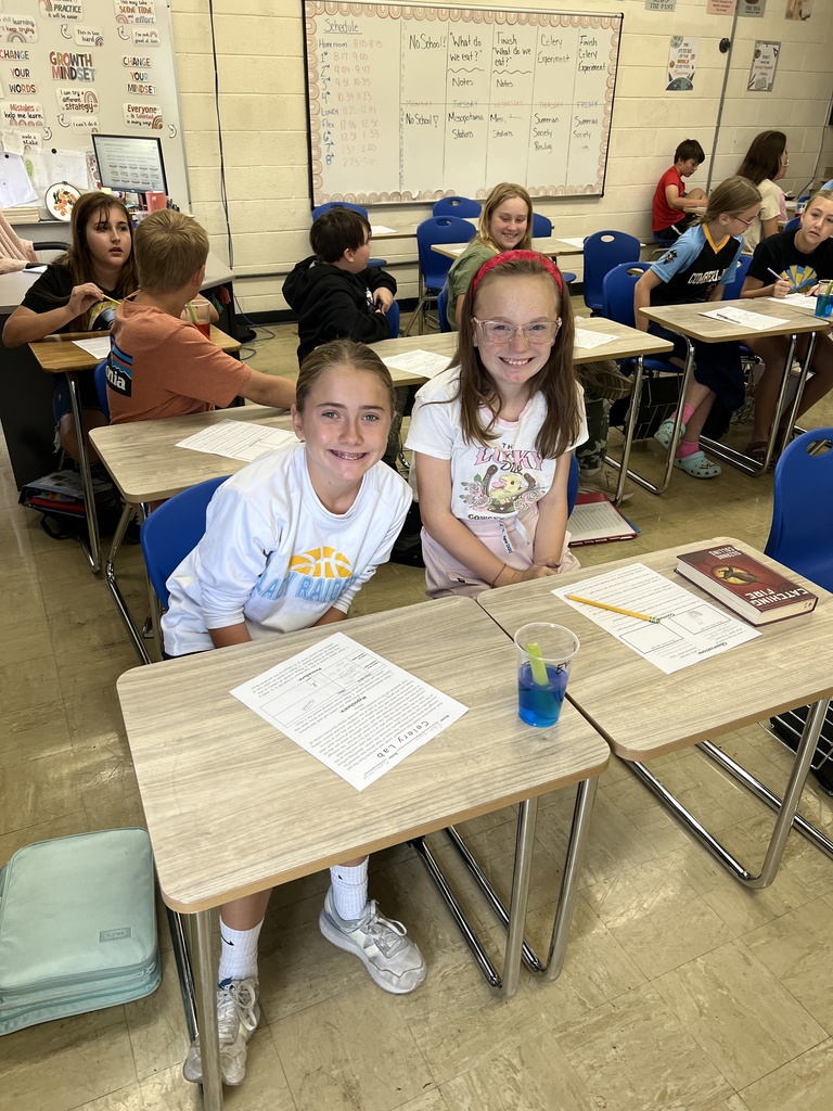 Two students are smiling for the camera. They are observing a celery stalk in a cup of blue dyed water. 