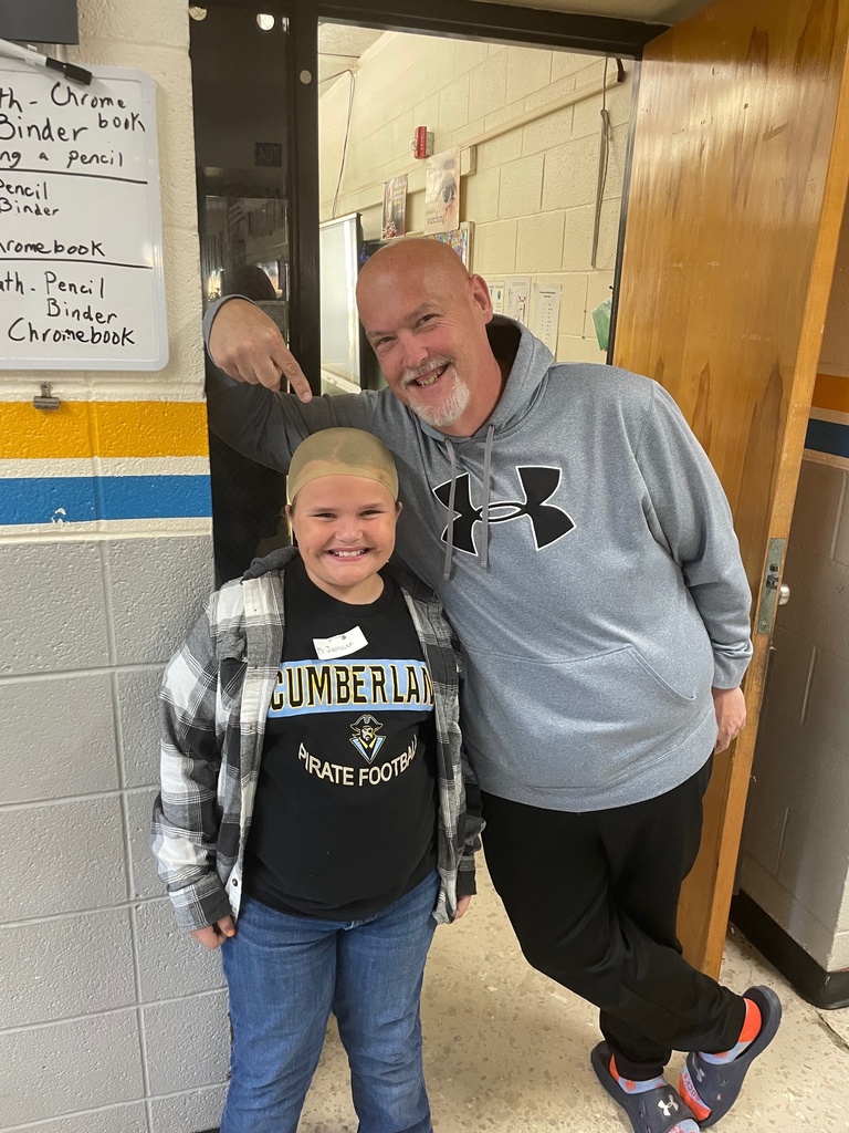 A student is standing wearing a bald cap. The teacher is wearing sweats, socks, and slides. The teacher is pointing to the bald head.