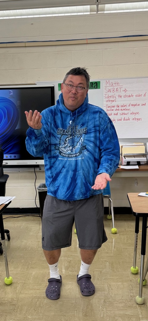 The teacher has gym shorts, a sweatshirt, socks and slides. He is guestering like the students do.
