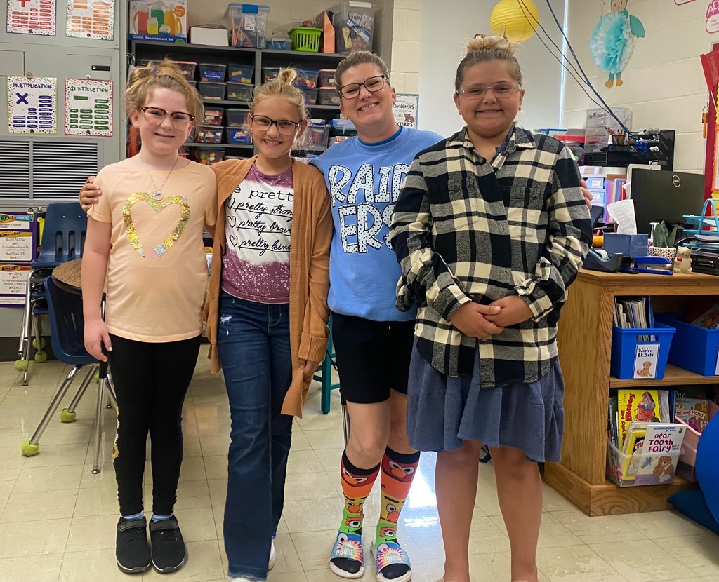 Three students are dressed as their teacher They are wearing dress clothers and have their hair in a bun. The teacher has a sweatshirt, shorts, crazy socks, and slides.