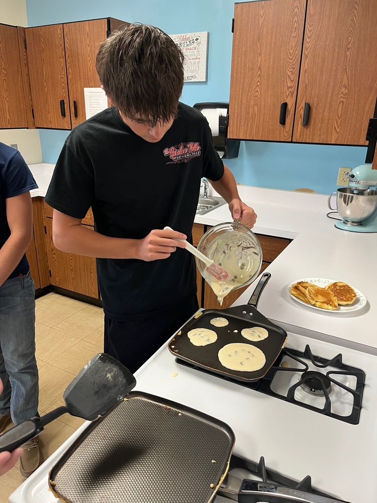 A student is standing in front of a stove. He is holding a class container with batter. He is pouring pancakes.