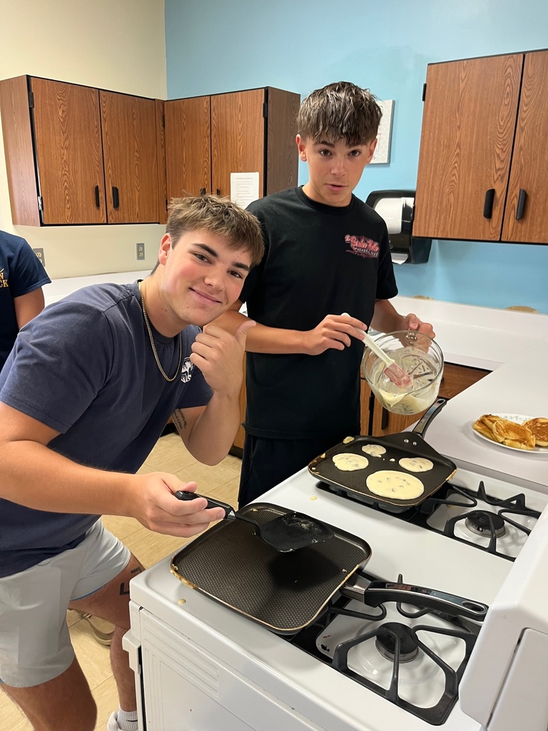 One student is pouring pancake batter. A second student is holding a spatula and giving the thumbs up sign.