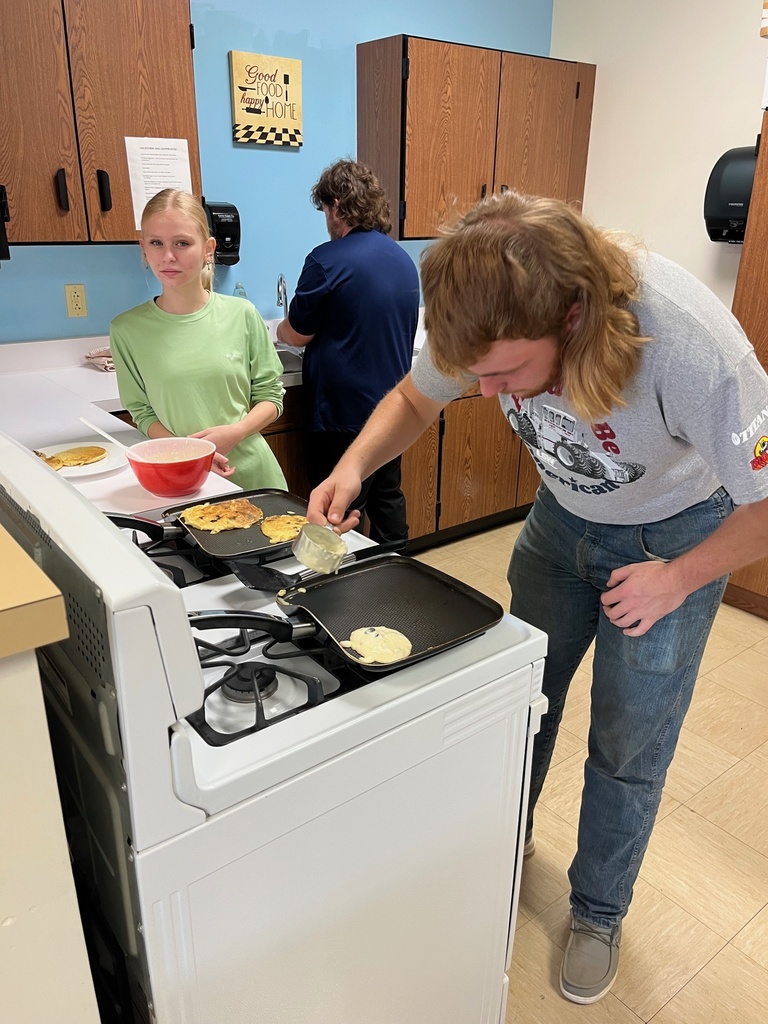 One student is standing in front of a stove holding a spatual over his pancake. Another student is leaning on the counter and watching.