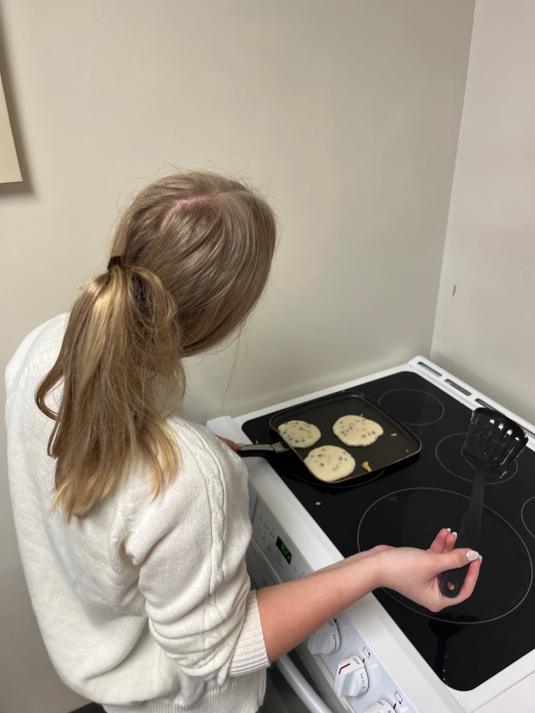 A student is standing in front of a stove. She is holding a spatula and watching her pancakes.