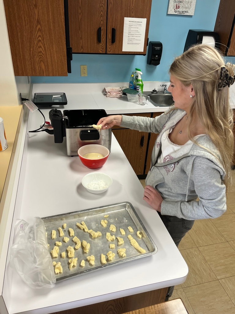 A student is standing at the counter. She is dipping cheese in batter and then holding it above the fryer.