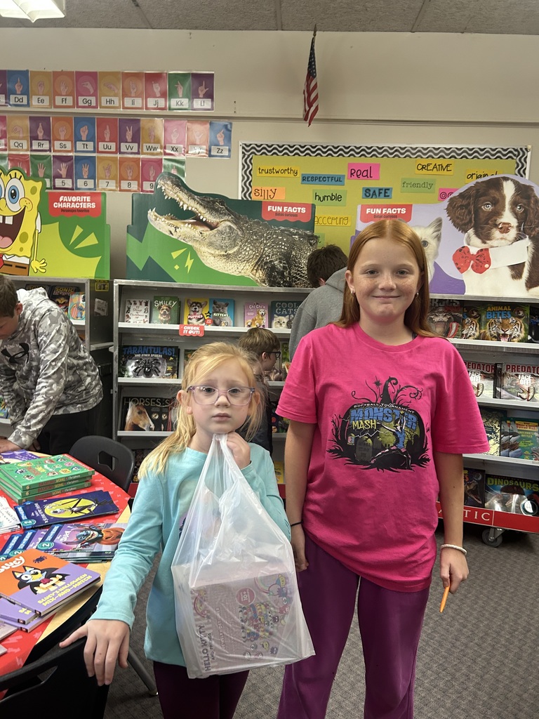 A yonger student is holding a bag of books and an older student is standing with her. They are at the book fair.