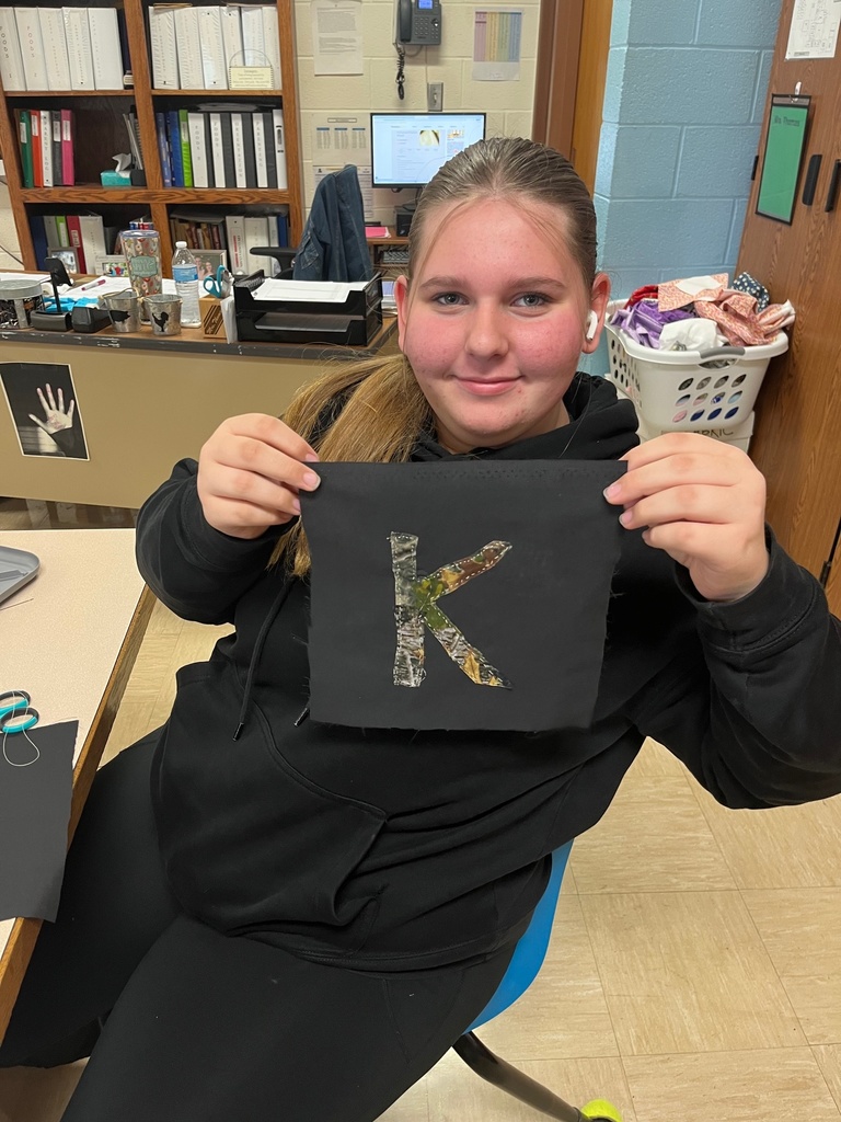 A student is sitting at a table. She is holding up a brown textile that has a camoflauge K sewn on it.