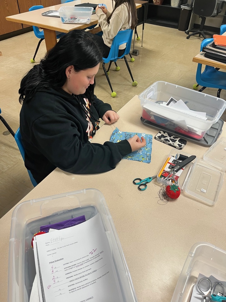 A student is sitting at a table. She is tying thread to the corner of a blue textile.