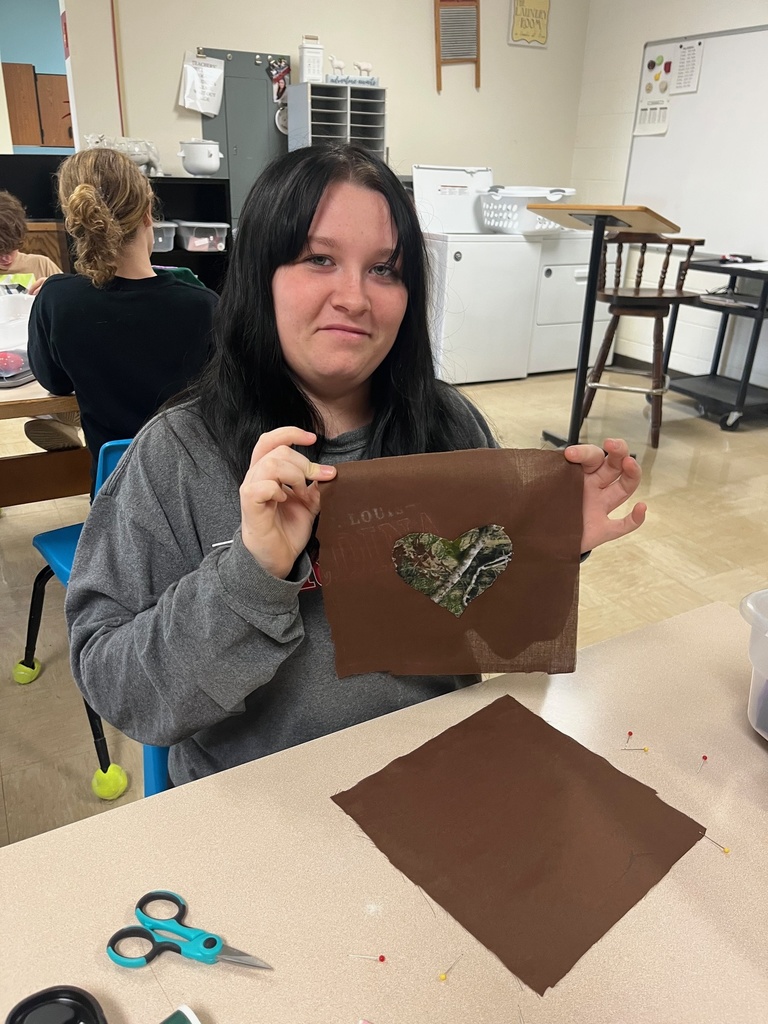 A student is sitting at a table. She is holding up a brown textile that has a camoflauge heart sewn on it.