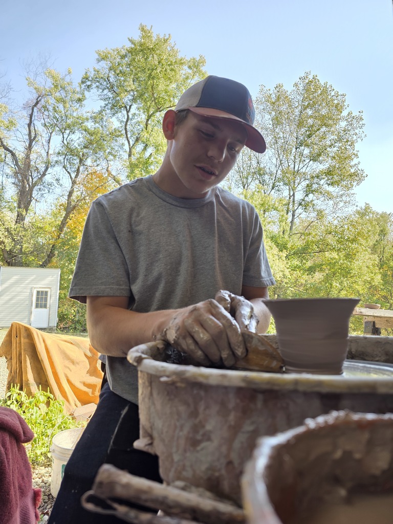 A student is at a throwing wheel. He is shaping the bottom of a ceramic dish.