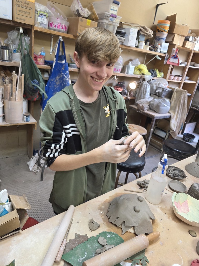 A student is standing at a table. He is molding clay to add to his creation on the desk.
