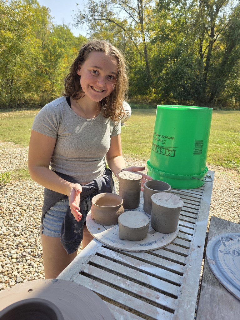 A student is standing outside. She has added her pottery to the round plate holding five clay projects.