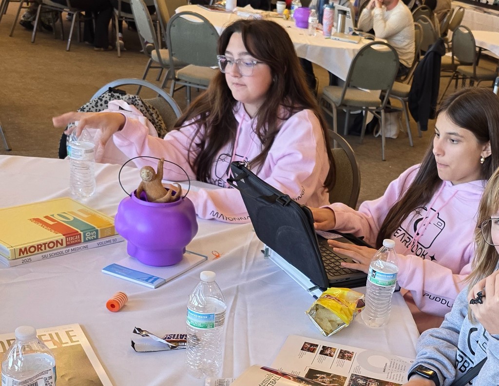 Two students is sitting at a table. One is working on a computer.