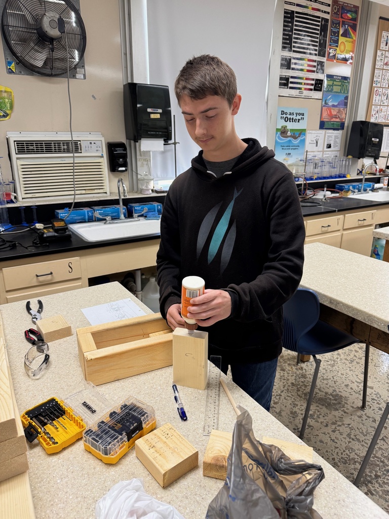 A student is standing at a table. He has wood in front of him. He is gluing the wood.