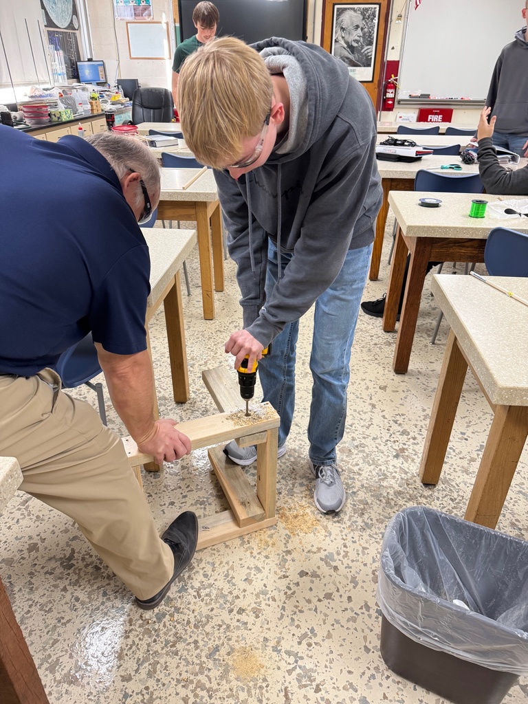 A student is drilling into wood. A teacher is stablizing the wood.
