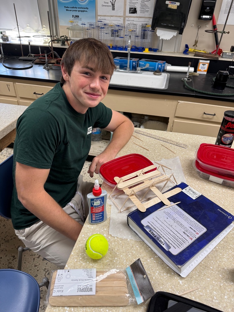 A student is sitting at a table. He has wood, glue, and a tennis ball in front of him.