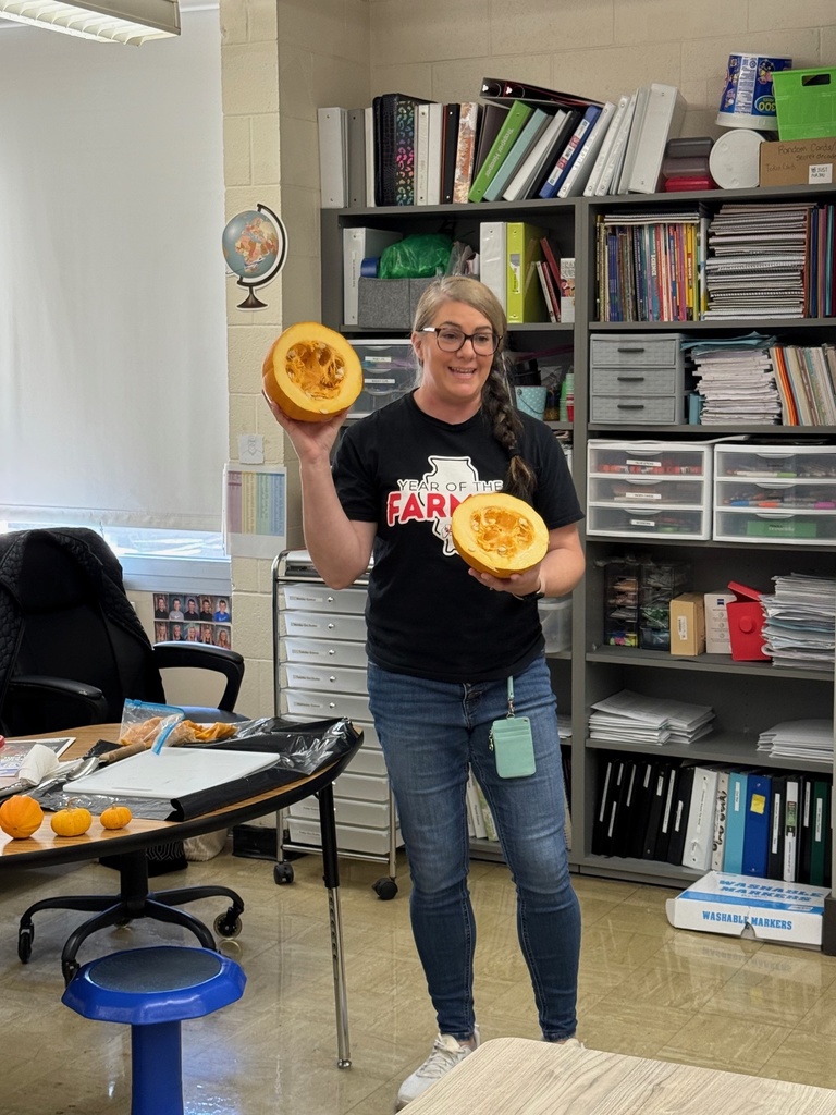 The teacher is standing in a classroom. She is hoding up two halves of a pumpkin.