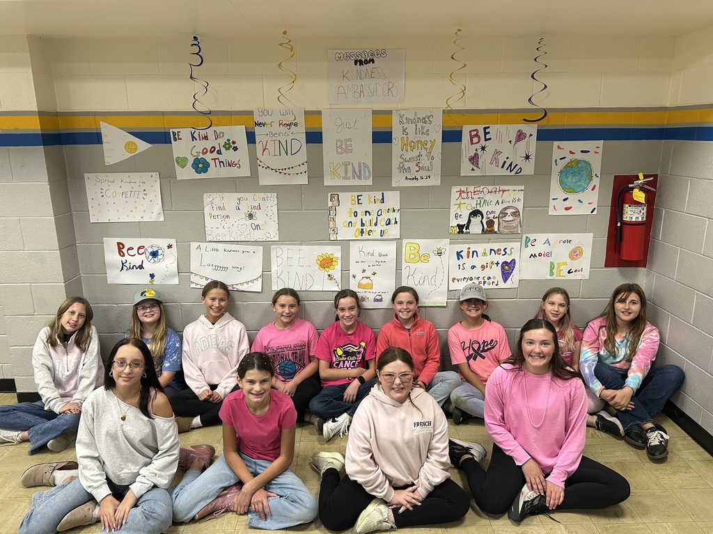 The Kindness Ambassadors are sitting on the floor in front of the wall they decorated. The wall has several posters hung with kindness messages.