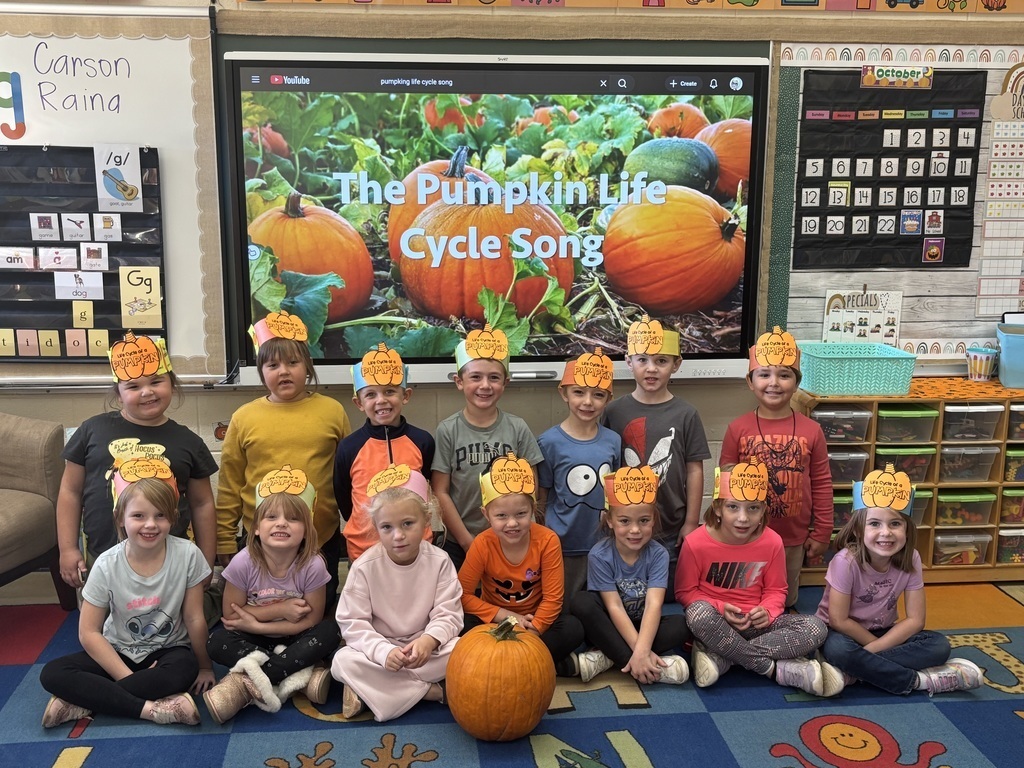 The class is sitting on the floor in front of a board that says The Pumpkin Life Cycle Song. The class is each wearing a pumpkin hat.