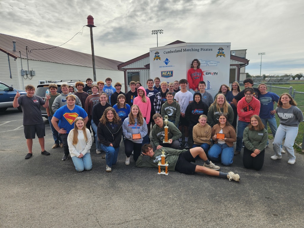 The band is standing together in front of their trailer. They are holidng their trophies and plaques from their competition season.