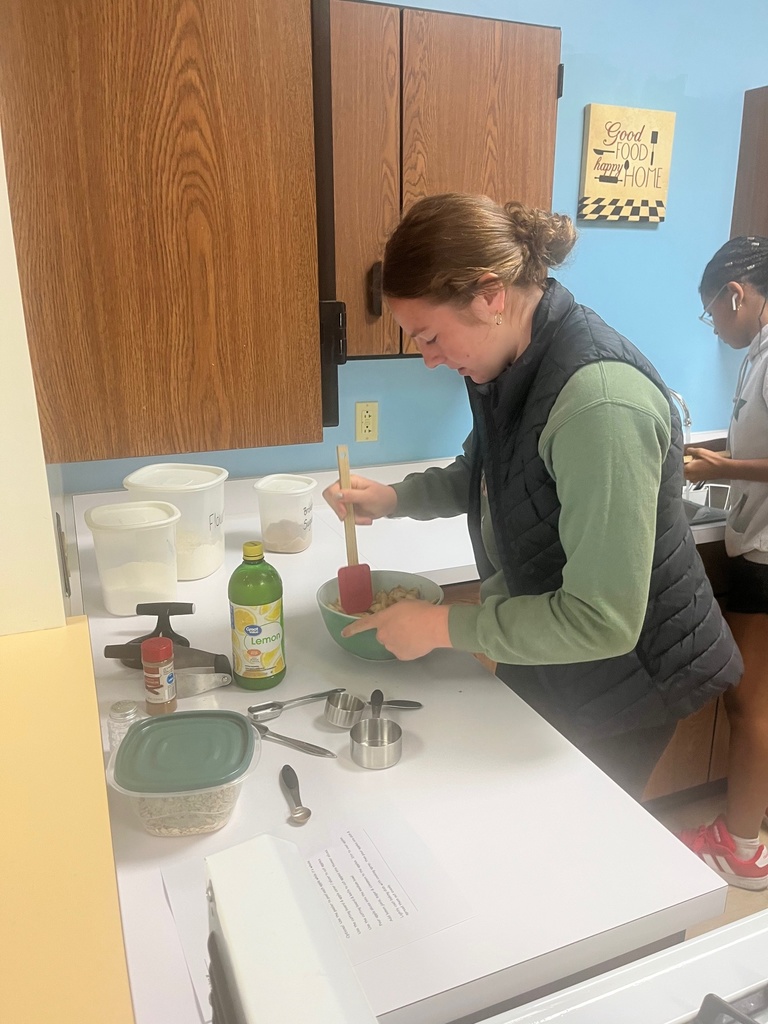 A student is standing at a counter. She is holding a mixing bowl and is stiring with a spatula.