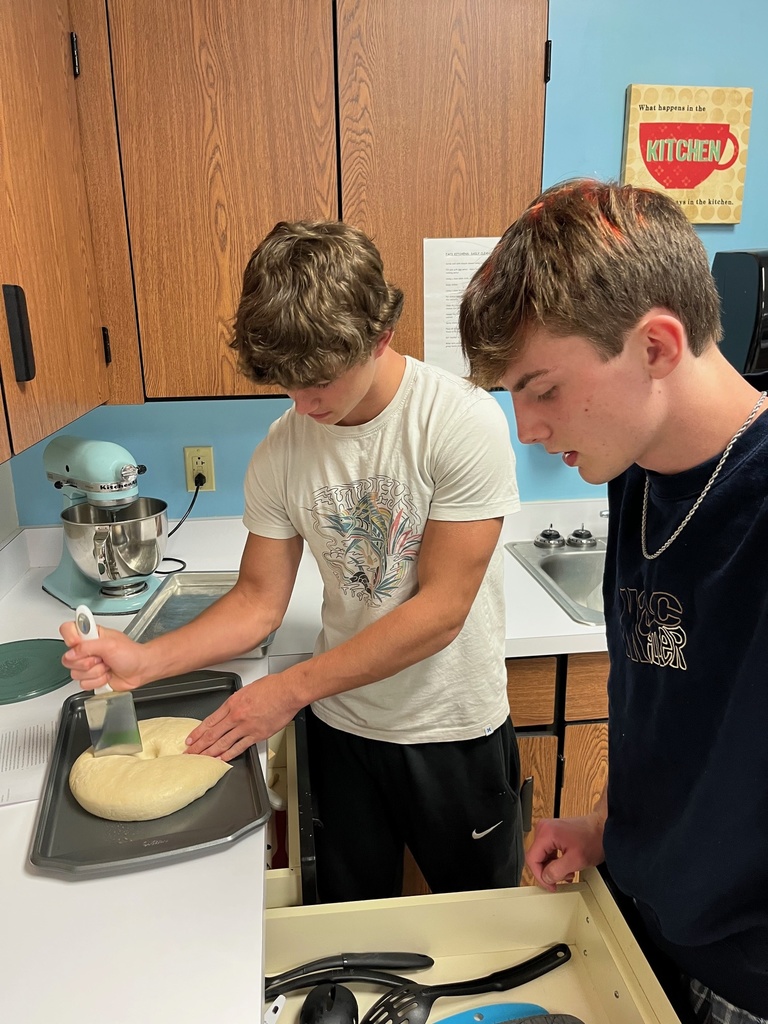 One student has the dough on a pan and is cutting it wit a spatula. The other student is looking in a drawer for baking supplies.